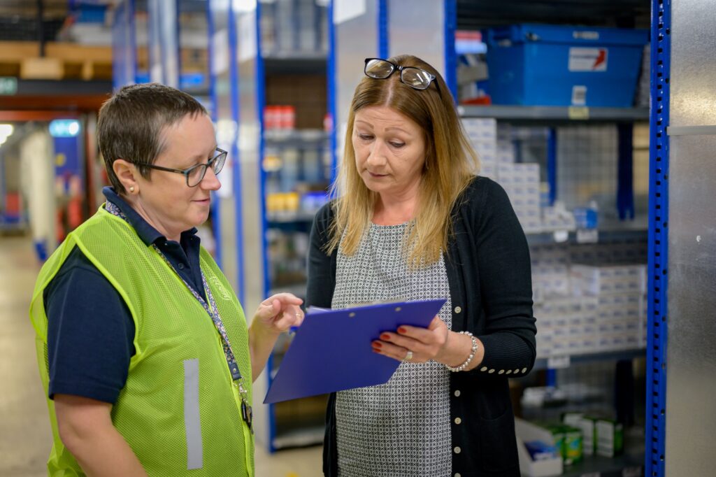 Two coworkers in a warehouse reviewing an inventory checklist together near storage shelving, an example of the peer coaching and knowledge-sharing that round out a balanced workplace training program