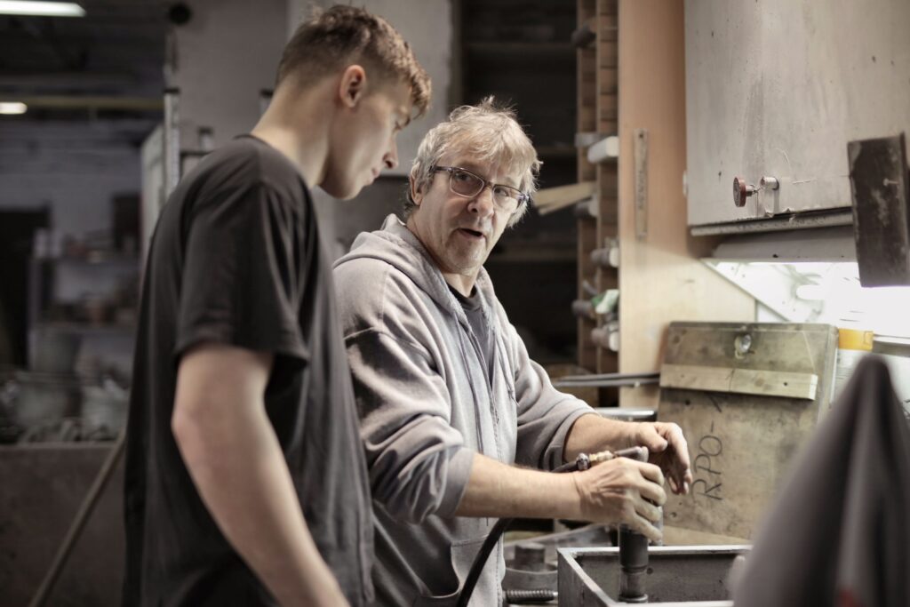 Experienced craftsman demonstrating pneumatic tool technique to a younger apprentice in a workshop, turning subject matter expertise into hands-on training content