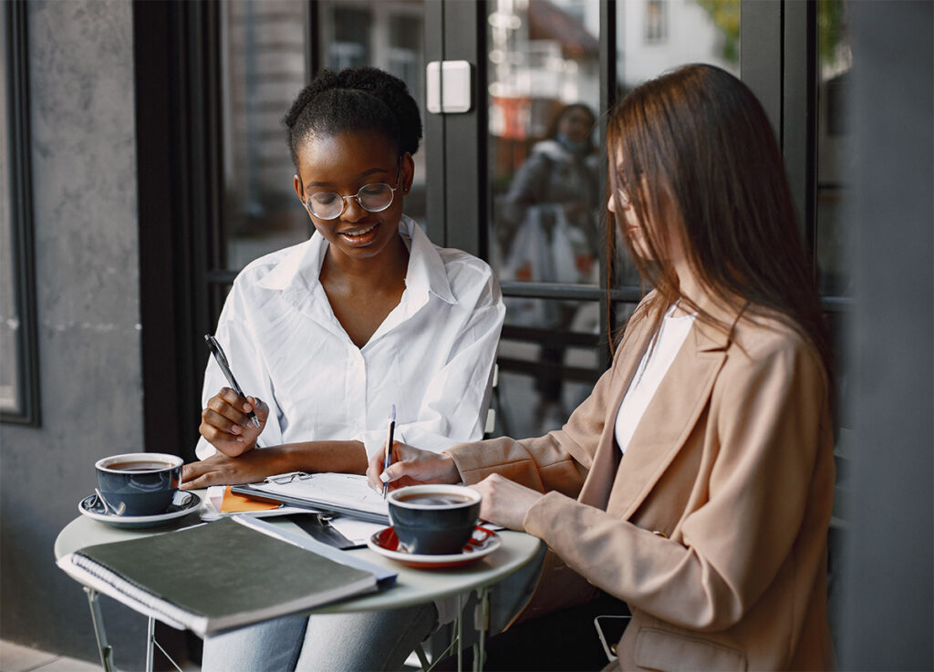 Two colleagues discussing a training needs analysis over coffee, one pointing out a detail on a shared notebook while the other looks on